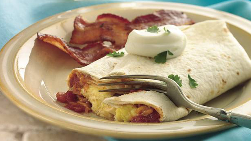 breakfast burritos topped with mayo served on a cream coloured plate with bacon